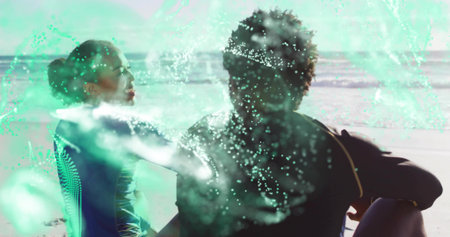 Sitting couple facing each other on beach near ocean, wearing wetsuits with green spray overlay....