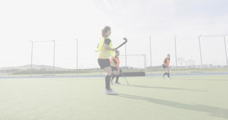 Holding hockey stick, woman in yellow bib wearing black shorts and socks on turf near goal. Scrimmage, practice, teammates, outdoor, turf, fencing, nettingの写真素材