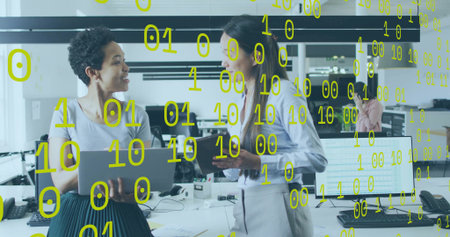 Talking two women in business attire gesturing over laptop at open office with yellow binary digits....