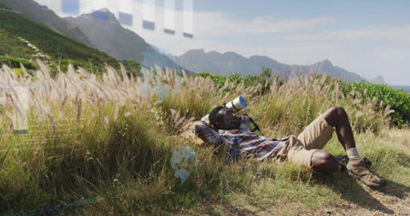 Lying hiker in plaid shirt khaki shorts boots, peering through scope at coastal meadow, bottle near....