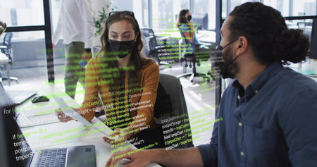 Working duo wearing masks leaning over laptop and papers in open office showing code overlay....