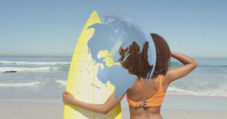 Holding yellow surfboard, woman in orange bikini top facing ocean on sandy beach, globe overlay....