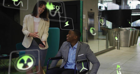 Discussing suited man in wheelchair and blazer-clad woman showing tablet at lobby with green icons. Collaboration, business, tech, presentation, modern, glass, loungeの写真素材