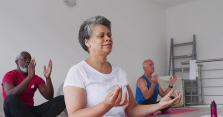 Sitting mature woman in white T-shirt breathing deeply in yoga studio, with mats and water bottle. Meditation, mindfulness, serenity, calm, focus, group, practiceの写真素材