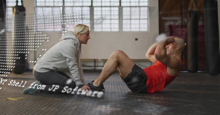 Coaching gray-hoodie trainer kneeling helping red-top trainee doing crunches on gym floor with bags. Boxing, training, duo, athlete, windows, daylight, industrialの写真素材