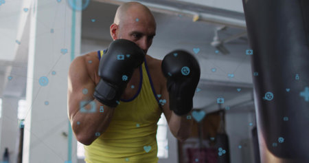 Training man with shaved head guarding punching bag at gym, wearing yellow top, black gloves, icons....