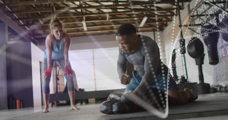 Leaning trainee in blue tank and red gloves watching kneeling trainer holding focus mitts in gym....