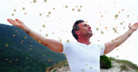 Standing mature man holding arms out on sand dune, wearing white t-shirt and golden sparkles....