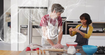 Baking couple wearing aprons kneading dough and cracking eggs at home kitchen island, flour cloud....