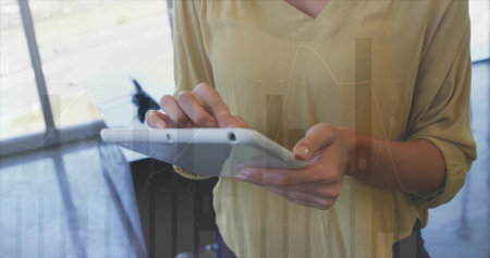 Holding white tablet, woman tapping screen in office near windows, wearing beige blouse with graphs....