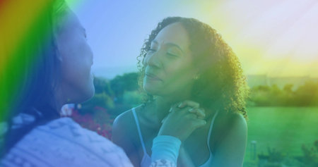Holding hands, two women wearing pale tank and patterned long-sleeve sitting in garden, lens flare. Duo, friendship, warmth, sunlit, greenspace, meadow, bokehの写真素材