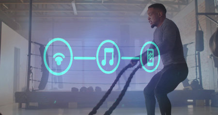 Swinging man in gray top powering battle ropes at boxing gym with glowing icons, copy space....