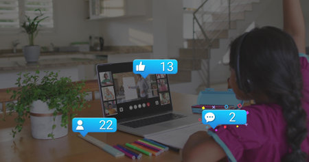 Sitting girl wearing pink top with blue trim and headphones raising hand at kitchen table, laptop. Kid, online, classroom, learning, daylight, stationery, greenery
