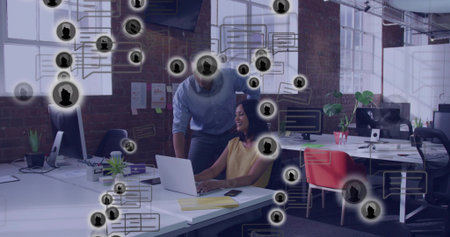 Leaning man in light shirt viewing laptop while woman in yellow blouse typing at office, overlays. Collaboration, desksharing, teamwork, workspace, openplan, daylight, brickwork