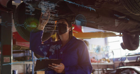 Inspecting tech in blue-coveralls and black-mask under lifted car in shop, holding tablet with data. Mechanic, garage, lift, diagnostics, augmented reality, overlay, inspection