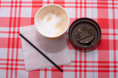 Yellow disposable glass of coffee with a straw and a napkin. Cardboard bench on a table with a red...