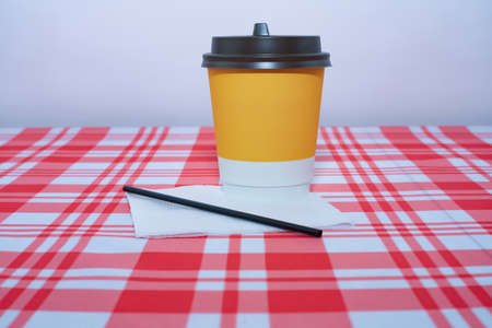 Yellow disposable glass of coffee with a straw and a napkin. Cardboard bench on a table with a red and white tablecloth. Fast food takeaway hot drinks packaging.の写真素材