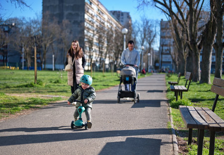 Little toddler on scooter and his parents with baby stroller walking in a parkの写真素材