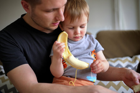 Father holding his little toddler helping him to peel bananaの写真素材