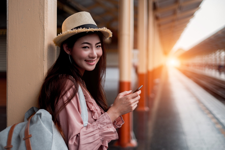 Smiling woman traveler looking camera with backpack holding smartphone on holiday relaxation at the train station,relaxation concept, travel conceptの写真素材