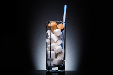 Glass with straw full of sugar and cubes on black background. The concept of prevention of diabetes, the harm of soda, overweight and dietの写真素材