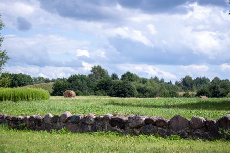 A large field with a stone wall and a hay bale in the middle. The hay bale is surrounded by grass and is placed in the center of the field. The field is open and has a peaceful, rural atmosphereの写真素材