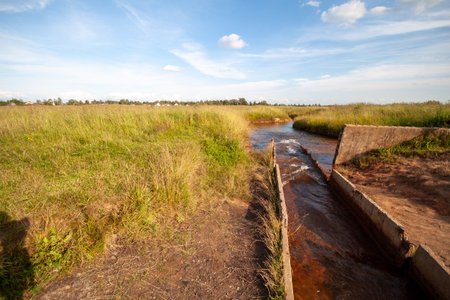 Water flow through a small concrete canal with metal gates and a field of grasses and shrubs in the background under a blue sky with white clouds.の写真素材