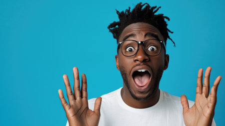 An African American male expresses surprise with wide eyes and open mouth against a blue studio background.の素材