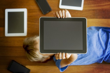 Little boy holding tablet overhead lying on wooden floor. Digital tablets, mobile phones, laptop all...