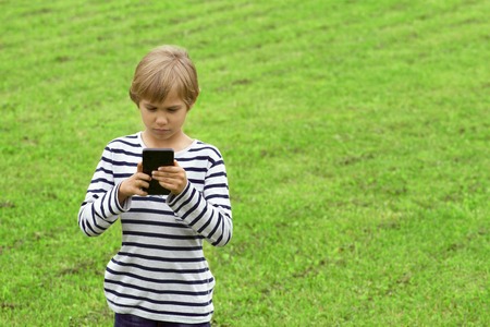 Boy with mobile smart phone outdoor. Child looking at the screen, playing, using apps. Green grass...