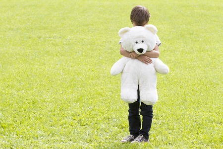 Disappointed boy is holding a teddy bear and standing on the meadow. Child looking down. Sadness fear frustration loneliness conceptの写真素材