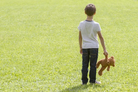 Sad young boy is holding a brown teddy bear and standing on the meadow. Back view. adness fear frustration loneliness conceptの写真素材