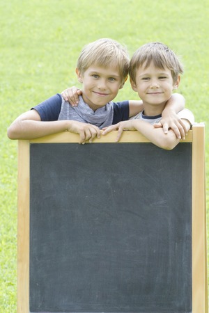 Two little boys standing at the blackboard outside. Green background. Copy space for text.の写真素材