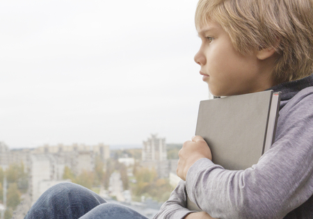 Boy with a book looking through the window. Child is concentrated, worried, sad, tired.の写真素材