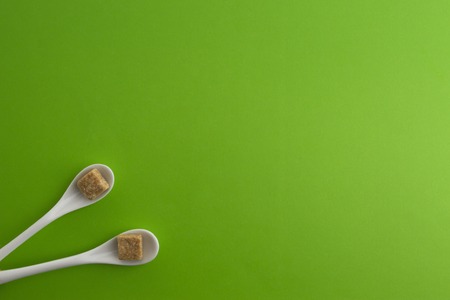 Two white spoons with brown sugar cubes placed in a corner on greenery background. Top view. Flat lay. Copy space for text. Diet unhealty sweet addiction conceptの写真素材