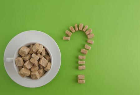 Diet, unhealthy sweet addiction concept. White cup full with brown sugar cubes and question mark of them on greenery background. Top view.の写真素材