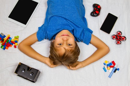 Cute smiling little boy lying in white bed and looking at camera. Mobile phone, tablet computer, drone and VR glasses around him.の写真素材