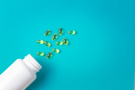 Omega 3 fish oil capsules spilling out of white plastic bottle on a blue background.