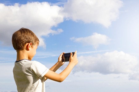 Boy using mobile phone. Child taking photo with his smartphone. Beautiful sky background. Back view. Technology conceptの写真素材
