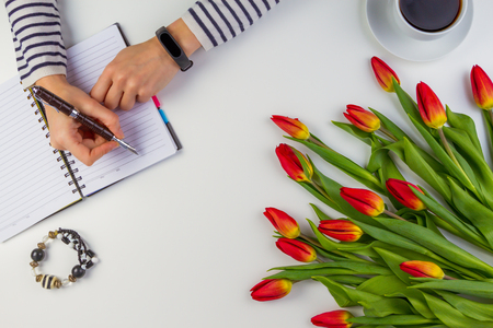 Woman hand writes on paper notebook with pen. Beautiful flowers on white table.の写真素材