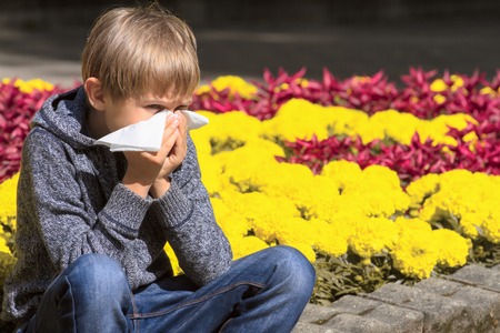 Child having allergy. Boy sitting outdoor with tissue near flowersの写真素材