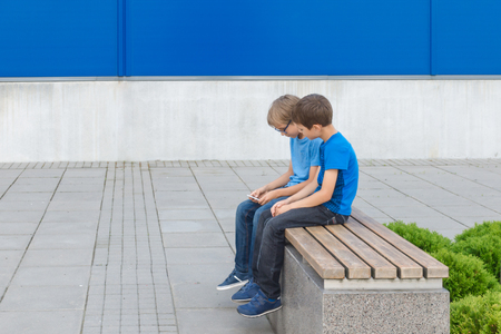 Two little boys playing with mobile phone sitting on the bench outdoorsの写真素材