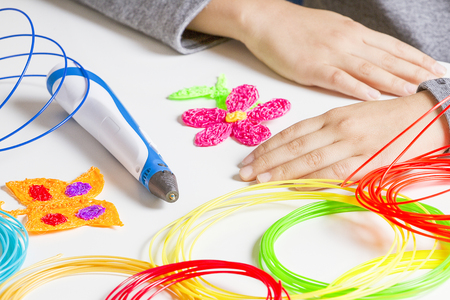 Kid hands and 3d printing pen, colorful filaments on white desk.