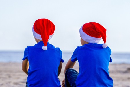 Kids in red Santa hats sitting at sunny beach during Christmas vacation and looking to seaの写真素材
