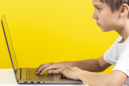 Kid sitting at the table and using laptop computer, yellow background