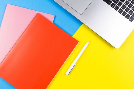 Top view of laptop computer, paper notebooks and white pen on blue and yellow color backgroundの写真素材