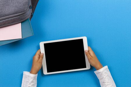 Top view to kid hands with tablet computer, backpack with books and notebook on light blue backgroundの写真素材