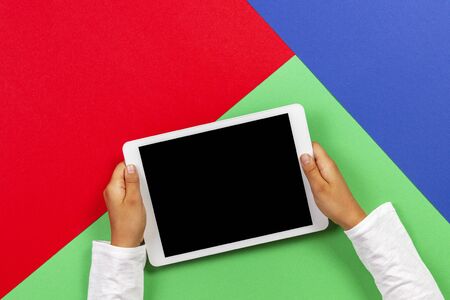 Kid hands holding white tablet computer on light green, blue and red backgroundの写真素材