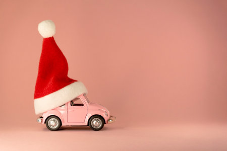 Christmas background. Little retro toy model car with small red Christmas Santa Claus hat on pastel pink backgroundのeditorial素材