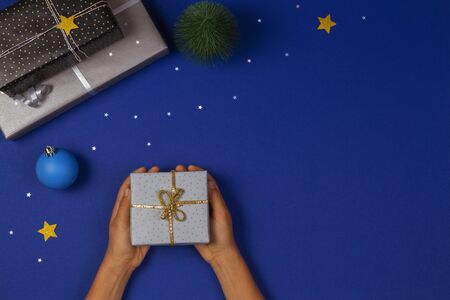 Christmas background. Top view to hands holding Christmas present box on navy blue background with sparkling glitter stars and bauble decorationの写真素材